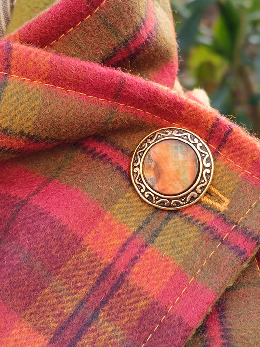 Détail du bouton vintage doré à motif marbré orange et crème, monté sur tartan en laine grattée rouge, rose, vert et jaune. Surpiqûres jaunes visibles. Accessoire textile artisanal mêlant pureté des lignes et esthétique rétro.