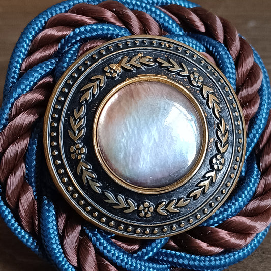 Broche ronde en passementerie avec cordon tressé bleu et brun. Au centre, un bouton vintage en métal ouvragé décoré de motifs floraux encadre un cabochon nacré irisé. La broche est posée sur une surface en bois, avec un élément sculpté flou en arrière‑plan. détaille des matériaux