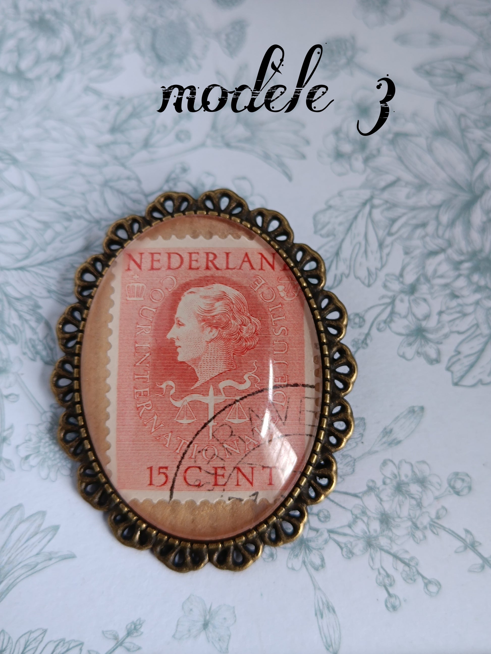 Broche ovale encadrant un timbre néerlandais vintage rouge du siècle dernier, illustré d’un profil féminin et marqué "NEDERLAND INTERNATIONALE JUSTITIE 15 CENT", avec oblitération partielle. Présentée sur un fond fleuri bleu pâle.
