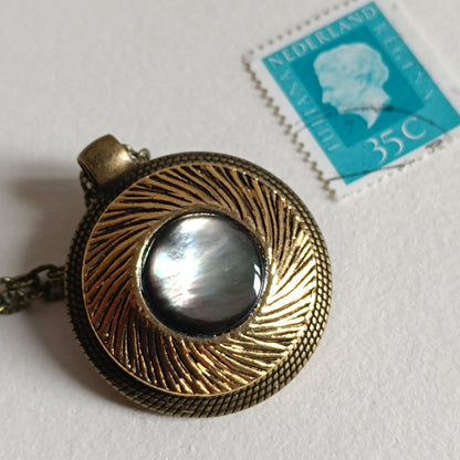 Pendentif vintage en bronze antique avec bouton ancien en nacre irisée, motif circulaire texturé, présenté à côté d’un timbre bleu pour l’échelle. Collier artisanal rétro années 50‑70.