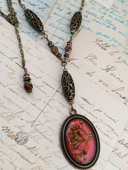Collier cottagecore artisanal en résine et fleurs séchées sur fond rouge, monture en bronze antique et perles de verre tchèque par Nathalie Aucoudesanges.