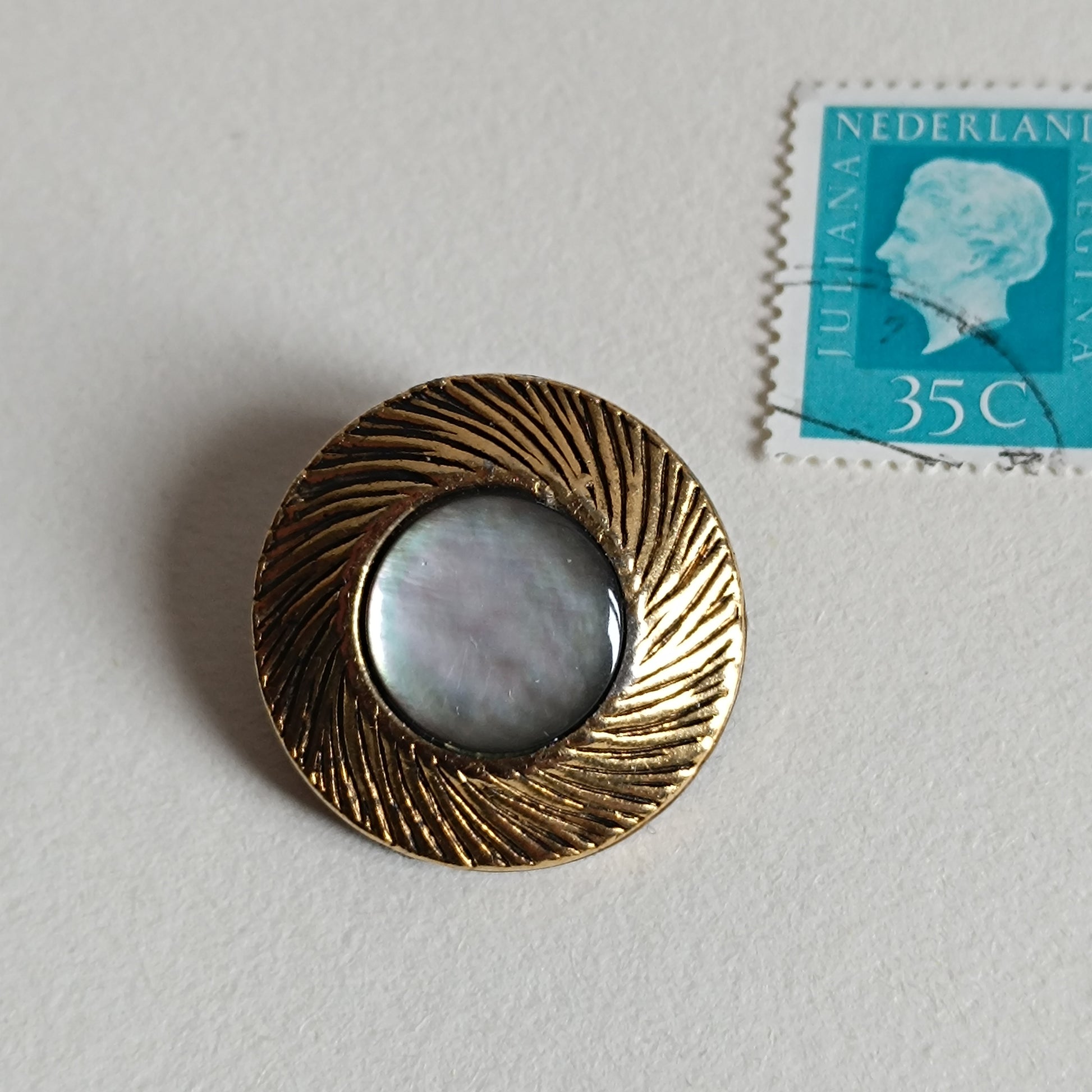Broche bouton ancien en métal doré texturé avec centre en nacre irisée, posée à côté d’un timbre vintage pour montrer la dimension. Accessoire rétro années 1950‑70.