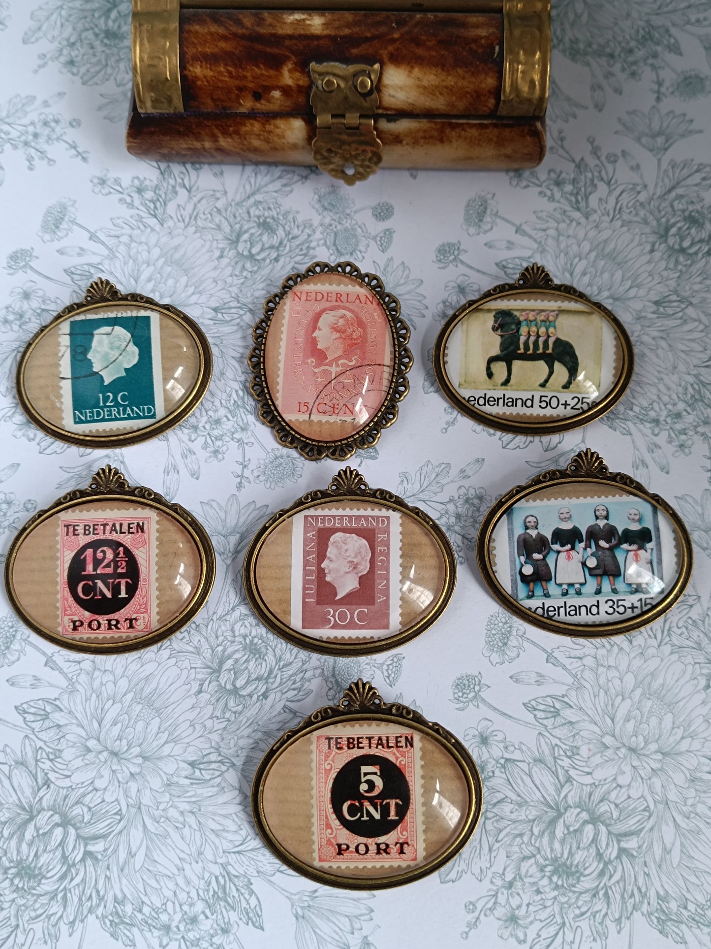 Collection de broches ovales encadrant de véritables timbres anciens néerlandais du siècle dernier, disposées sur un fond fleuri avec un petit coffret en laiton. Chaque timbre présente une illustration différente – portrait royal, typographie d’époque ou motif culturel – évoquant l’univers de la philatélie vintage.