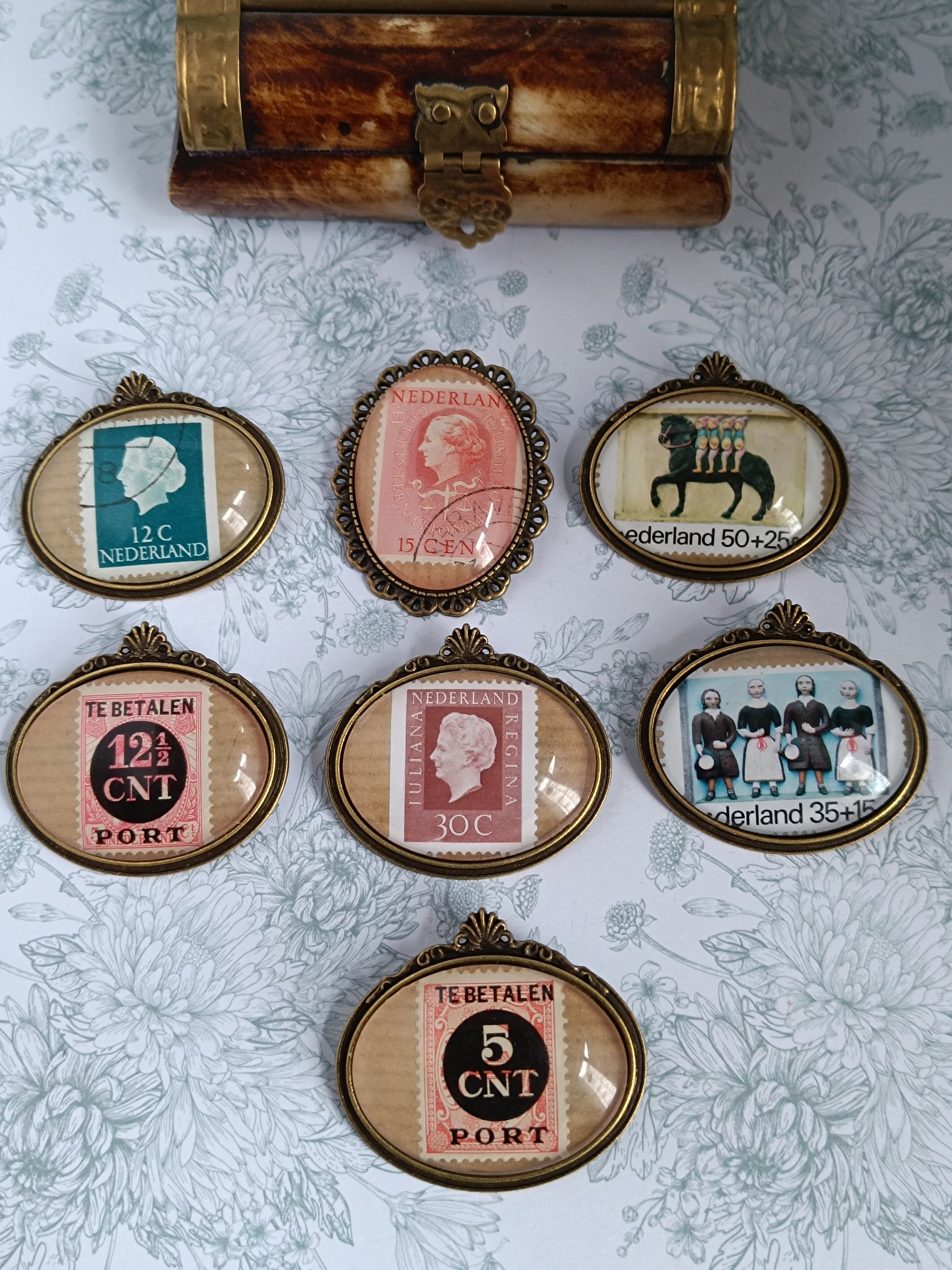 Collection de broches ovales encadrant de véritables timbres anciens néerlandais du siècle dernier, disposées sur un fond fleuri avec un petit coffret en laiton. Chaque timbre présente une illustration différente – portrait royal, typographie d’époque ou motif culturel – évoquant l’univers de la philatélie vintage.