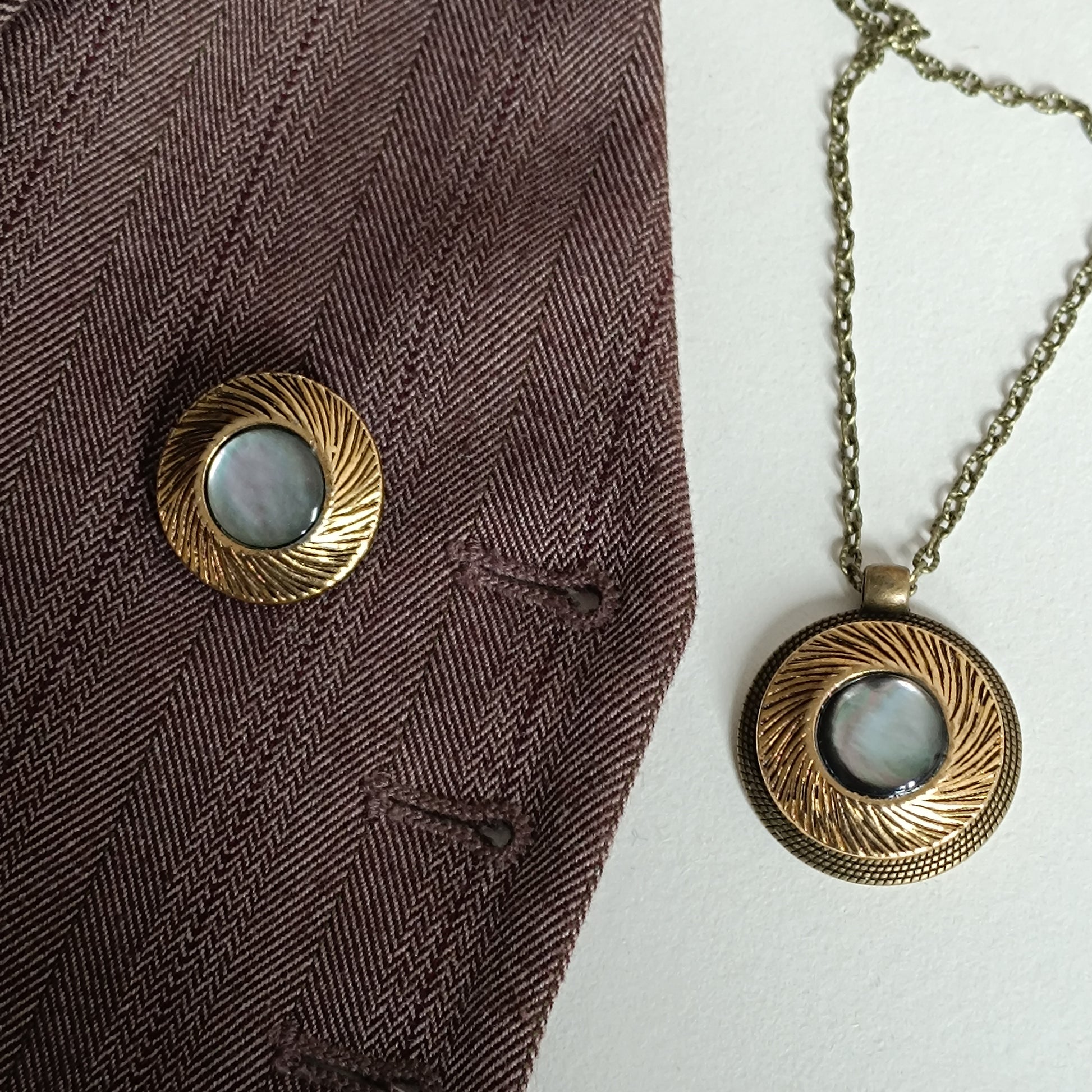 Pendentif vintage en nacre irisée et métal doré présenté sur tissu chevron marron foncé pour montrer l’effet du bijou sur vêtements sombres. Bouton ancien transformé en bijou artisanal, style rétro élégant et discret.