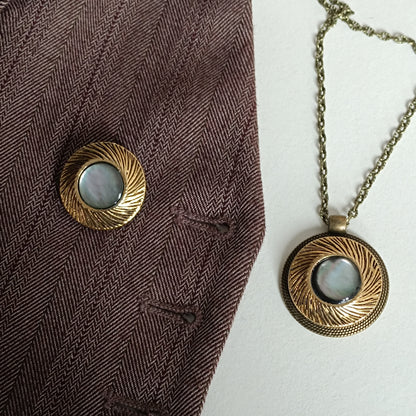 Pendentif vintage en nacre irisée et métal doré présenté sur tissu chevron marron foncé pour montrer l’effet du bijou sur vêtements sombres. Bouton ancien transformé en bijou artisanal, style rétro élégant et discret.
