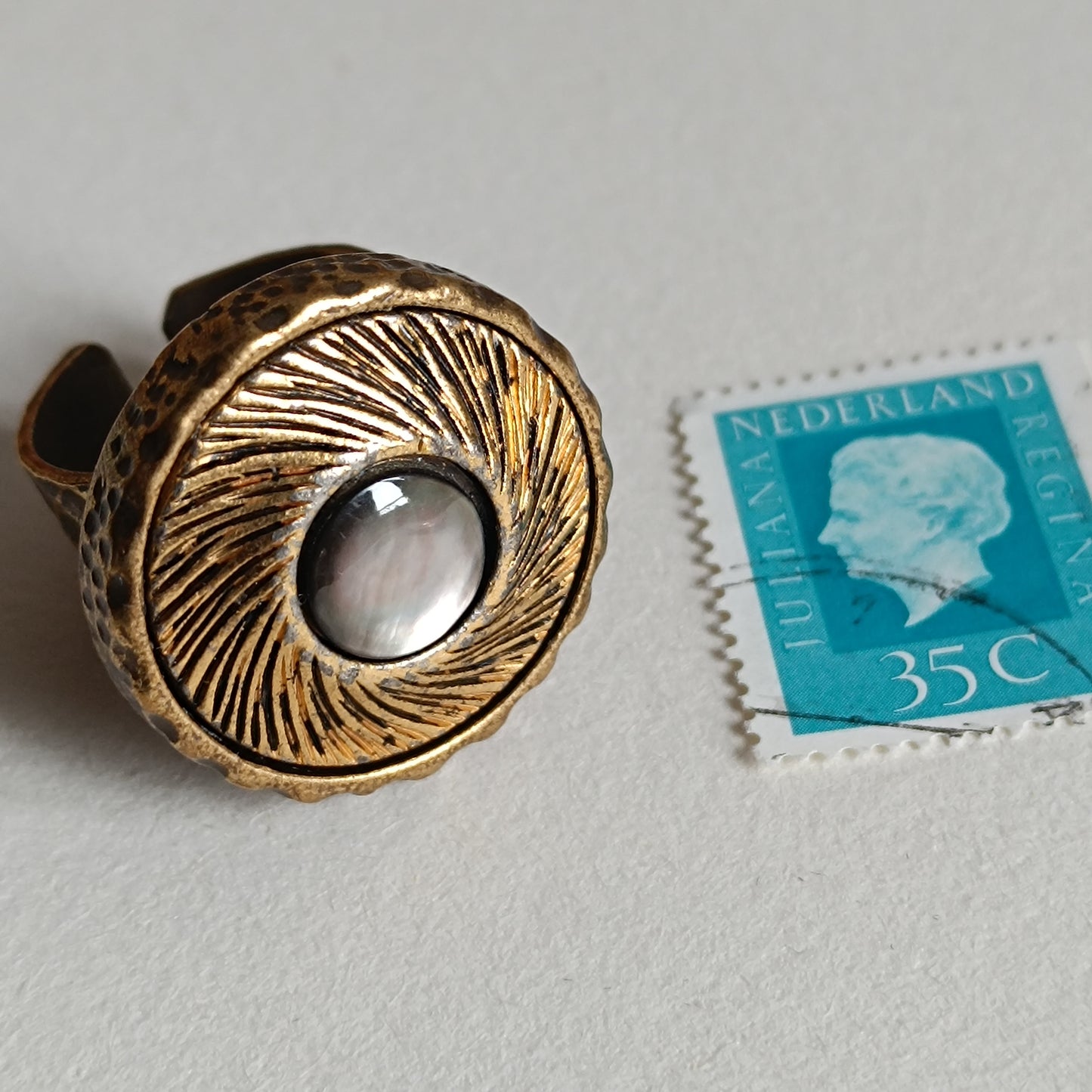Bague vintage en bronze antique avec large cabochon rond en nacre irisée, posée à côté d’un timbre ancien pour montrer l’échelle. Bijou artisanal rétro inspiré des boutons anciens.