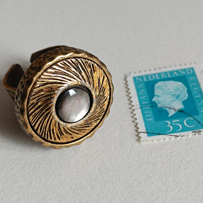 Bague vintage en bronze antique avec large cabochon rond en nacre irisée, posée à côté d’un timbre ancien pour montrer l’échelle. Bijou artisanal rétro inspiré des boutons anciens.