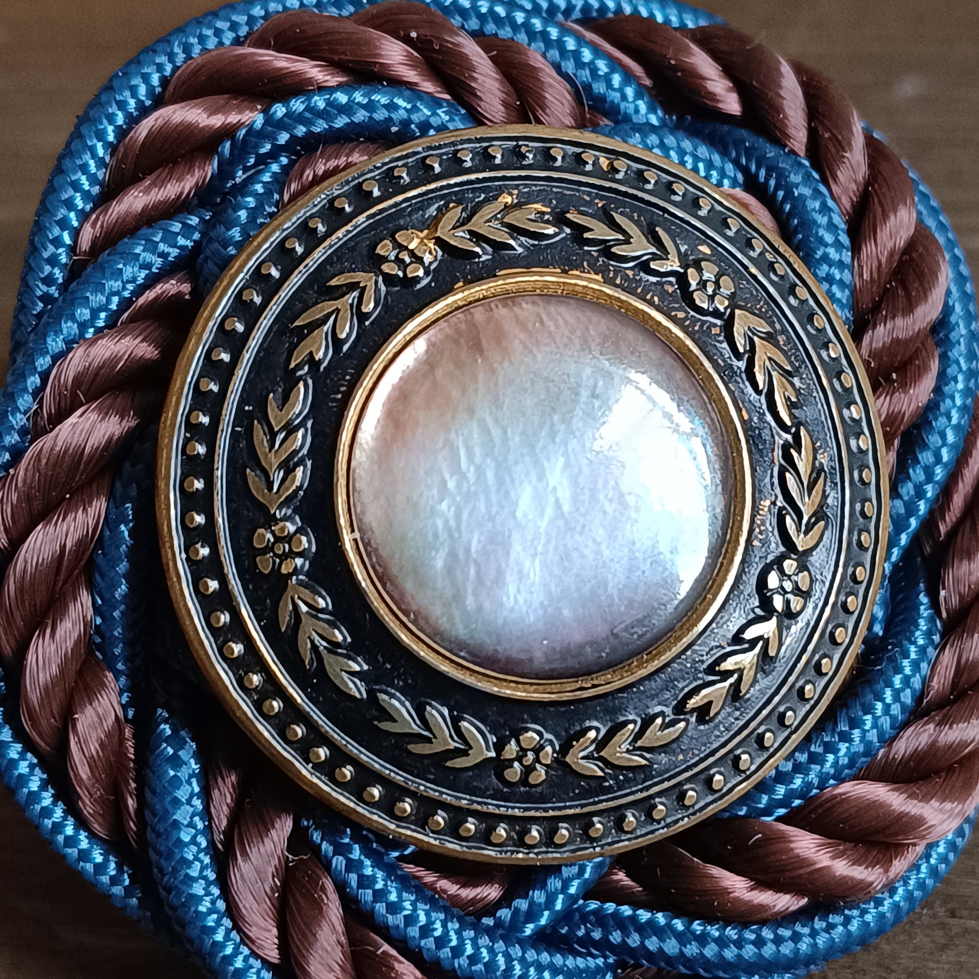 Broche ronde en passementerie avec cordon tressé bleu et brun. Au centre, un bouton vintage en métal ouvragé décoré de motifs floraux encadre un cabochon nacré irisé. La broche est posée sur une surface en bois, avec un élément sculpté flou en arrière‑plan. détaille des matériaux 