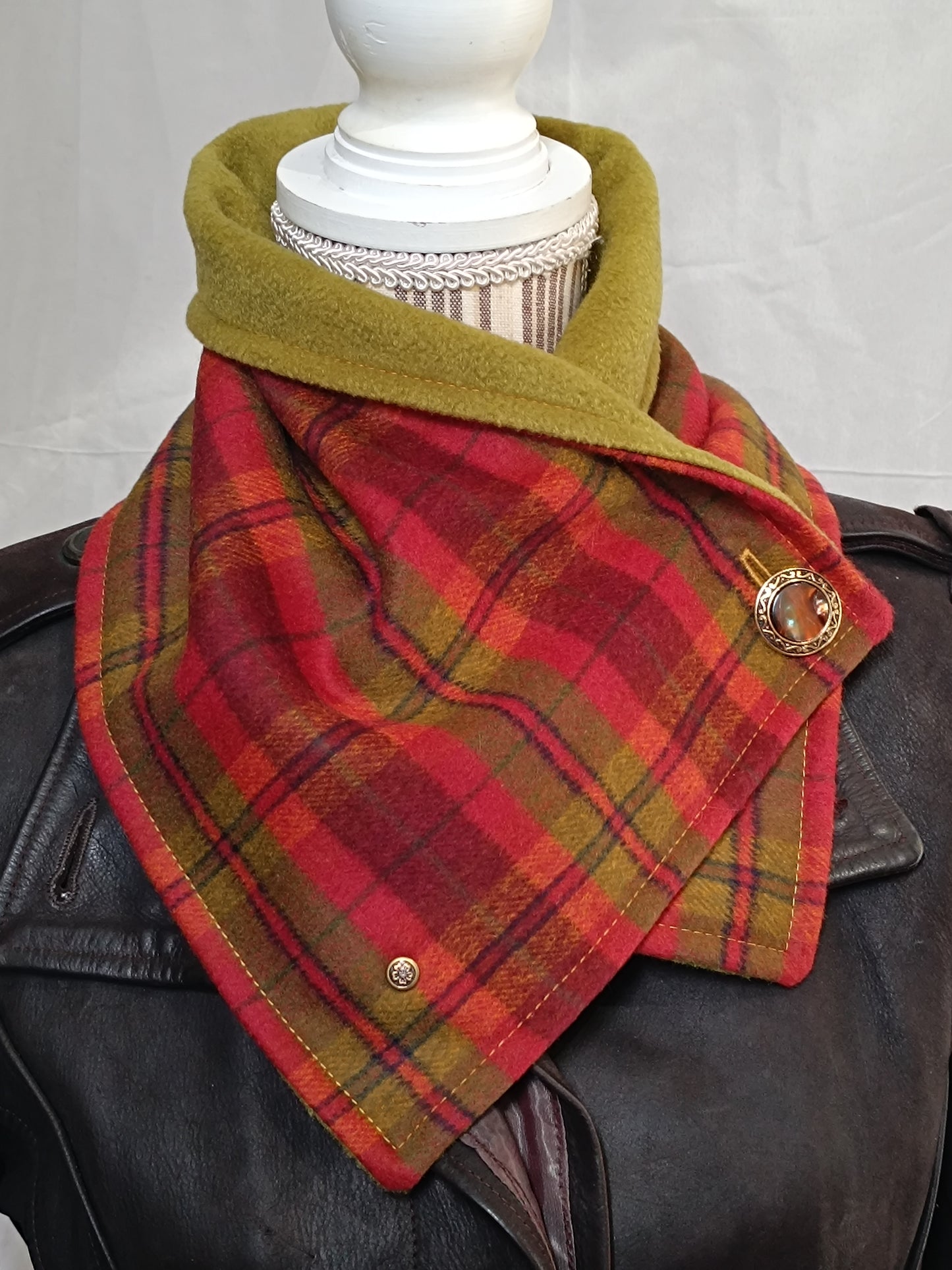 Tour de cou artisanal en laine grattée tartan rouge, vert et jaune doublé polaire vert pistache, porté sur veste en cuir foncé. Accessoire textile modulable : se porte ouvert façon col bénitier, fermé en ras-du-cou structuré, ou légèrement décalé pour un effet asymétrique affirmé. Bouton vintage doré et rivet bronze fleurs de cerisier visibles.