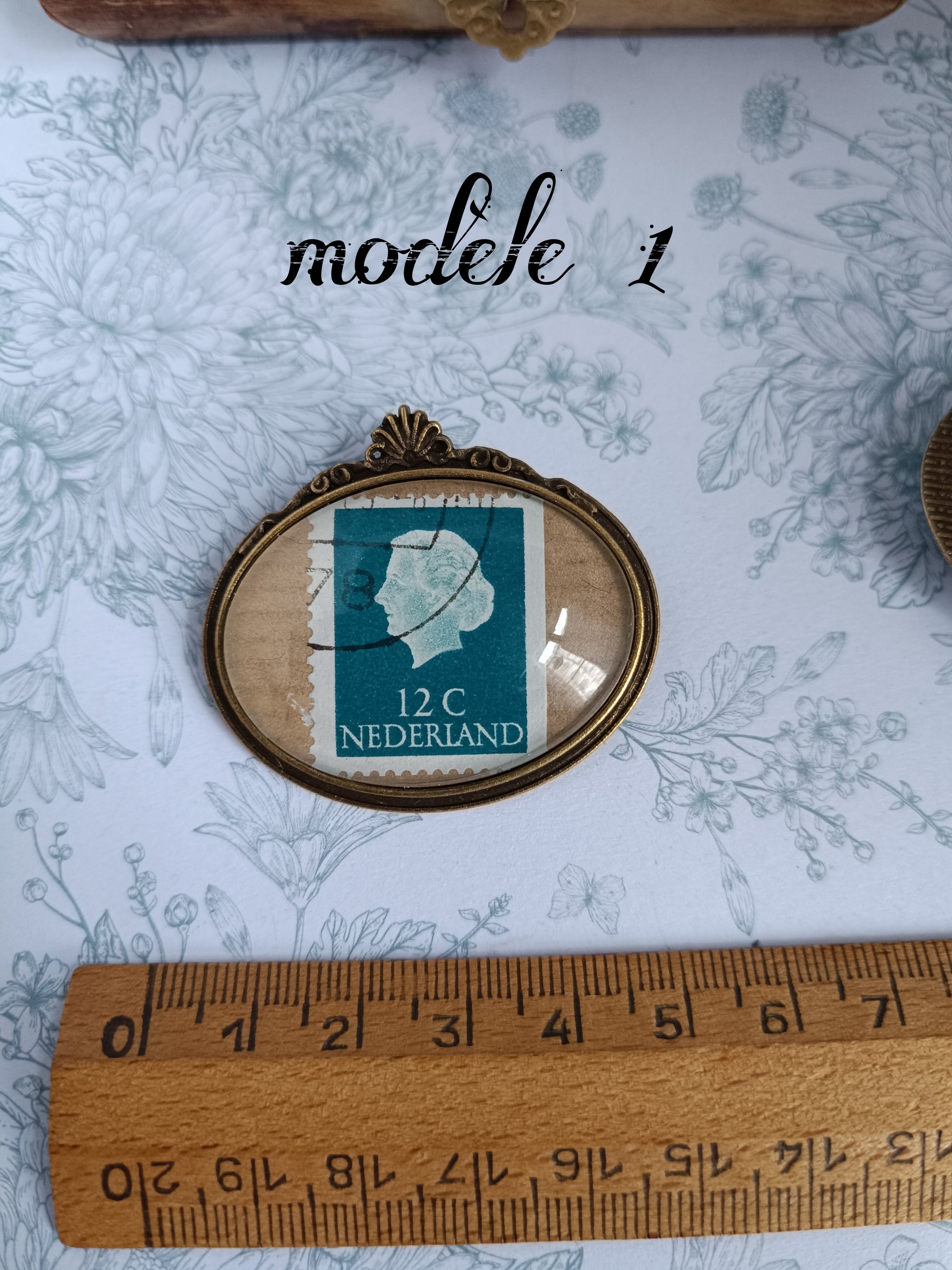 Broche ovale encadrant un timbre néerlandais vintage de 12 centimes, daté du siècle dernier, avec de la reine Régina profil blanc sur fond bleu-vert et marques d’oblitération visibles. Présentée sur un tissu fleuri avec une règle en bois indiquant une largeur d’environ 5 cm.