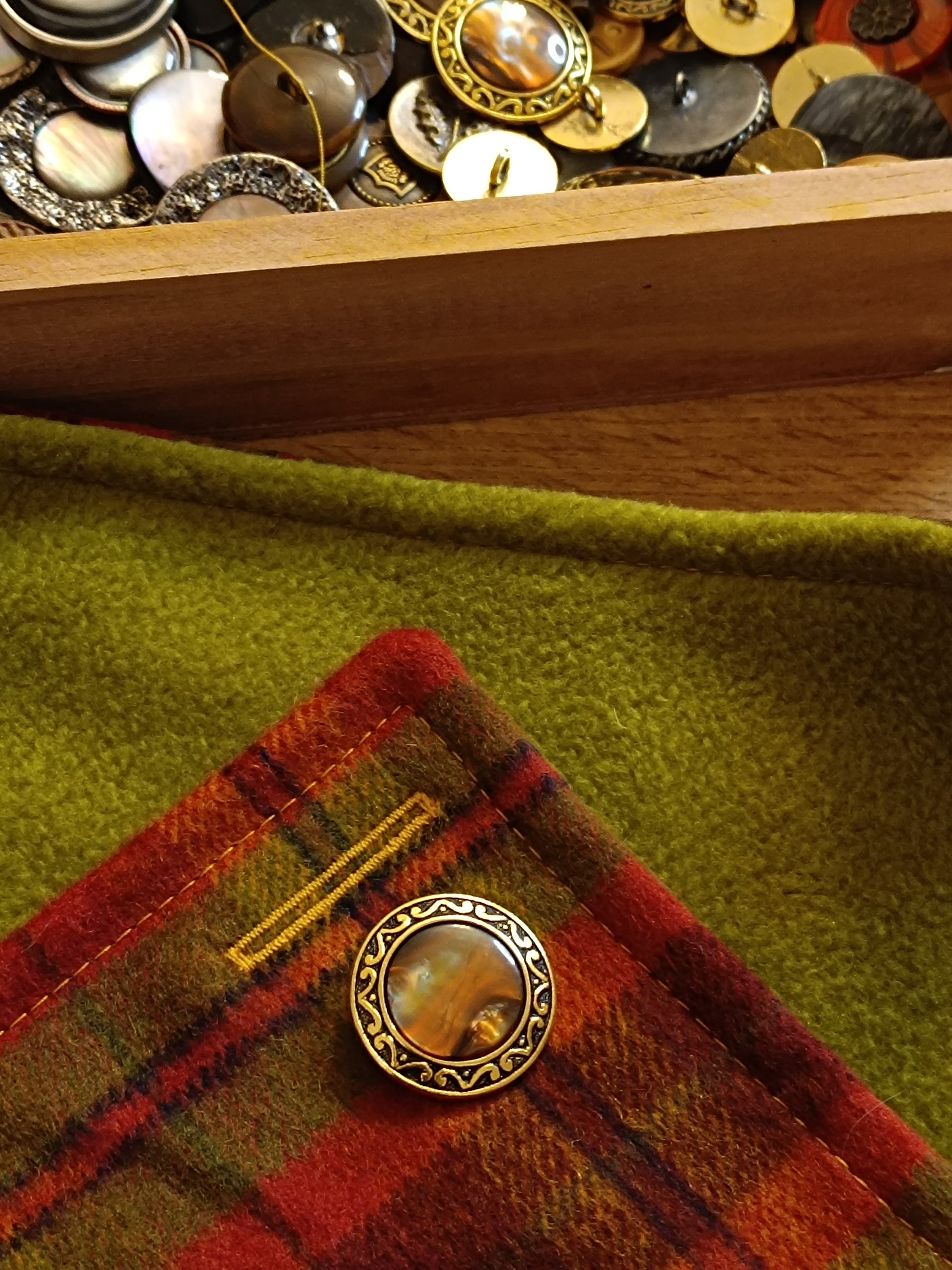 Boutonnière réalisée en fil jaune sur tartan en laine grattée rouge, orange, vert et jaune. Bouton vintage doré à centre marbré posé sur le tissu, en cours de confection. Photo d’atelier mettant en valeur le soin apporté aux finitions et le contraste lumineux des surpiqûres.