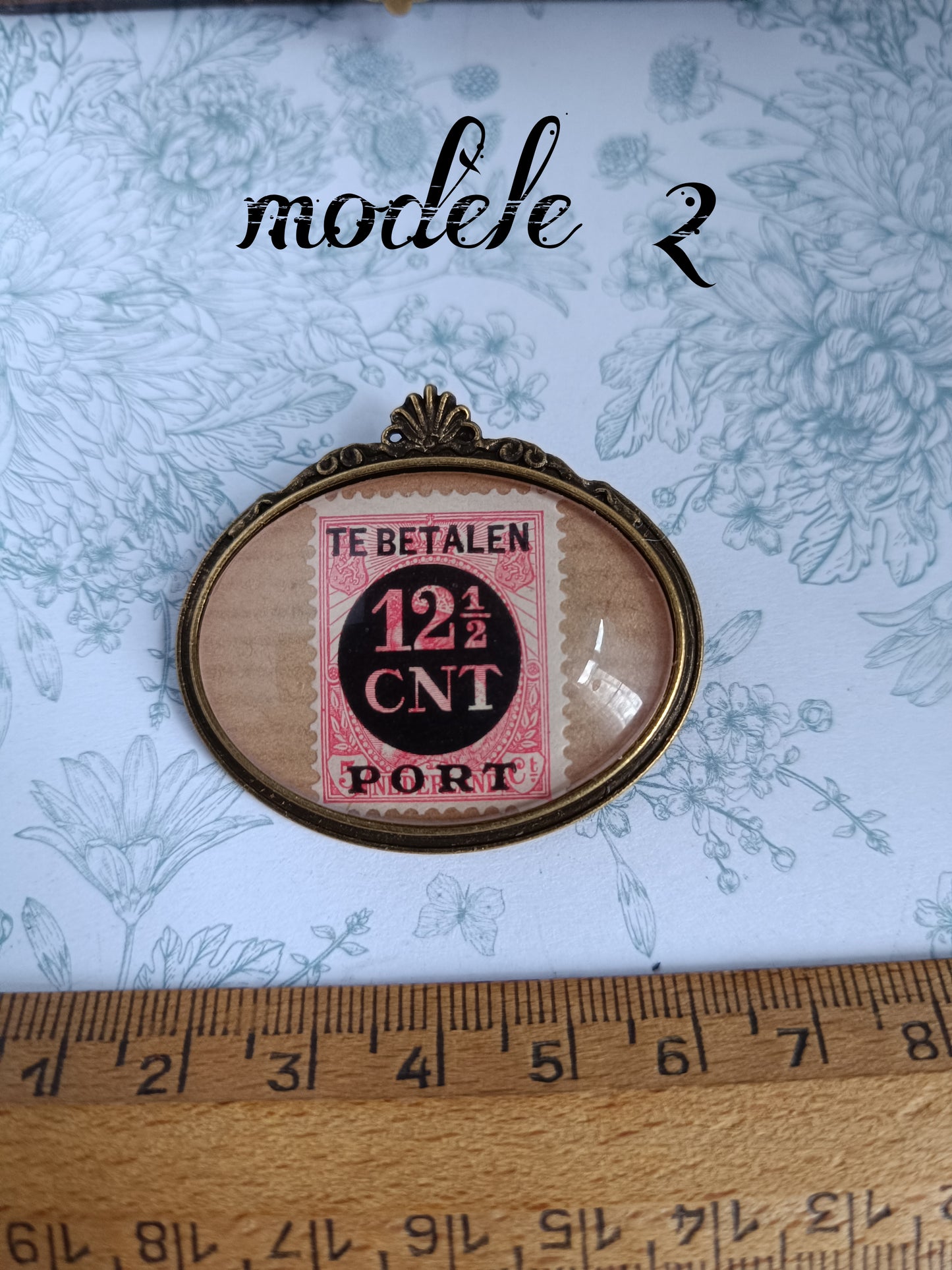 Broche ovale encadrant un timbre néerlandais vintage rose et noir du siècle dernier, marqué "TE BETALEN 12½ CNT PORT", avec oblitération visible. Présentée sur un tissu fleuri avec une règle en bois indiquant une largeur d’environ 1,5 pouce.