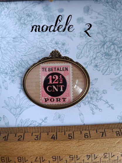 Broche ovale encadrant un timbre néerlandais vintage rose et noir du siècle dernier, marqué "TE BETALEN 12½ CNT PORT", avec oblitération visible. Présentée sur un tissu fleuri avec une règle en bois indiquant une largeur d’environ 1,5 pouce.