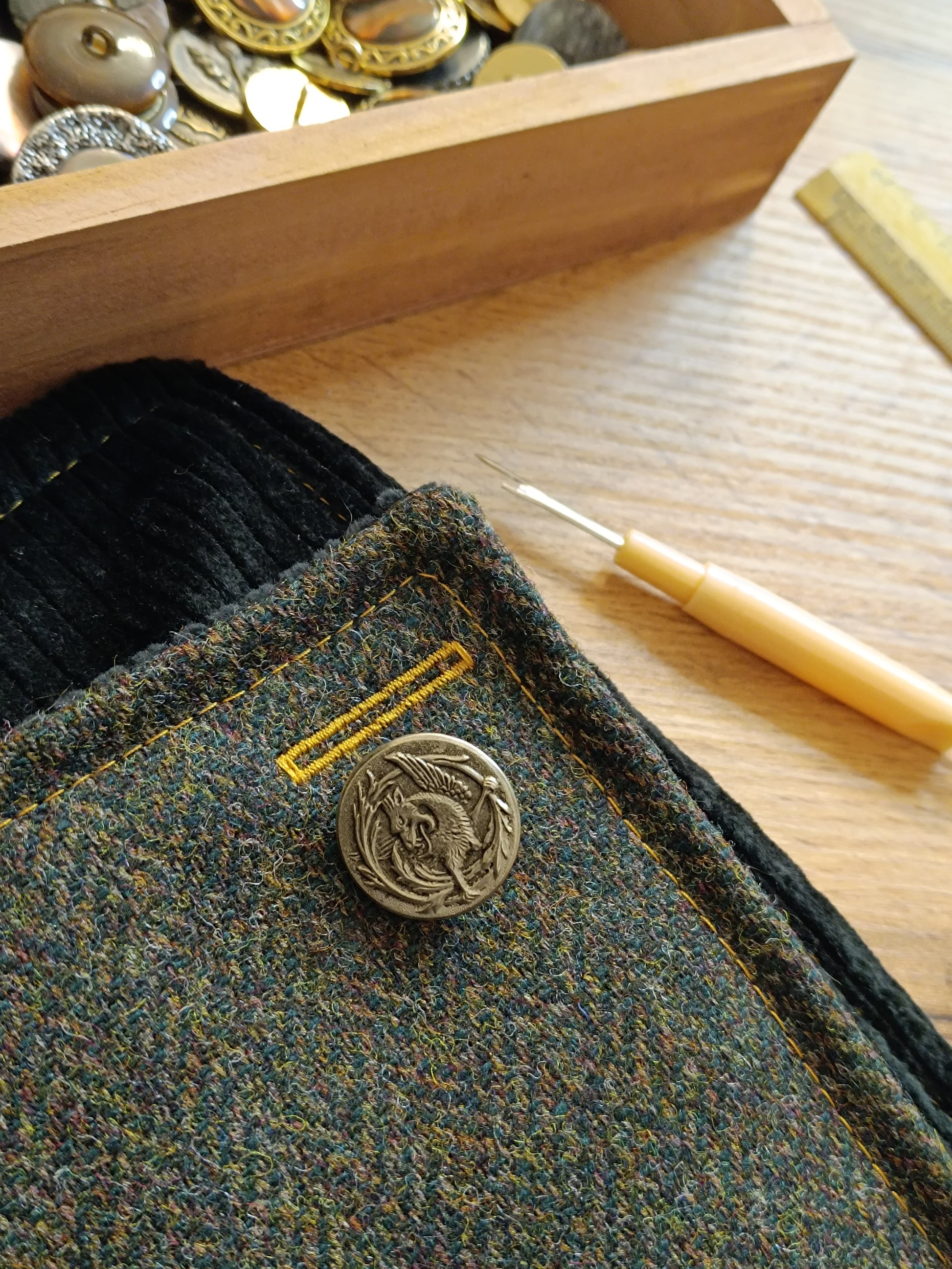 Bouton vintage en métal blanc représentant un dragon cracheur de feu, posé près d’une boutonnière cousue mais encore fermée. Surpiqûres jaune ocre sur tweed multicolore. Gros plan sur le travail en cours dans l’atelier : couture précise, matières choisies avec soin. Détail textile idéal pour accessoire fait main au style féerique, bohème ou médiéval, réalisé dans notre atelier de l’Oise.