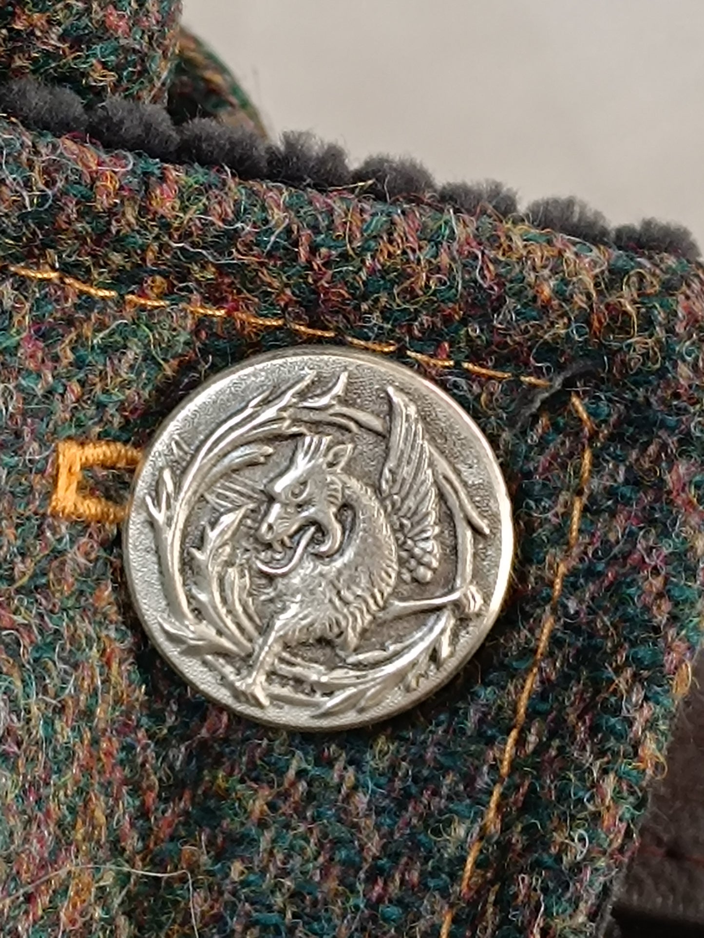 Bouton vintage en métal blanc représentant un dragon cracheur de feu, cerclé de feuillages stylisés. Fixé sur tweed multicolore aux reflets verts, bleus, rouges et jaunes, avec surpiqûres jaune ocre. Détail textile artisanal au style féerique, bohème ou médiéval, idéal pour accessoire fait main réalisé dans notre atelier de l’Oise.