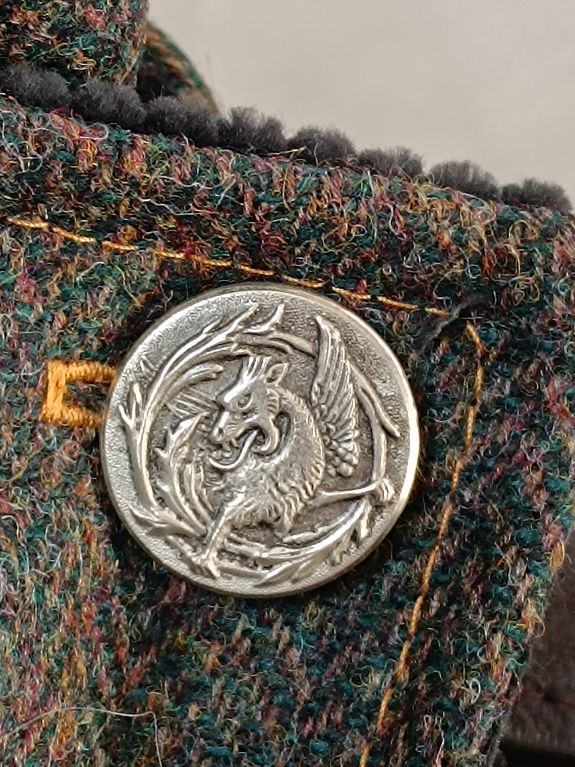 Bouton vintage en métal blanc représentant un dragon cracheur de feu, cerclé de feuillages stylisés. Fixé sur tweed multicolore aux reflets verts, bleus, rouges et jaunes, avec surpiqûres jaune ocre. Détail textile artisanal au style féerique, bohème ou médiéval, idéal pour accessoire fait main réalisé dans notre atelier de l’Oise.
