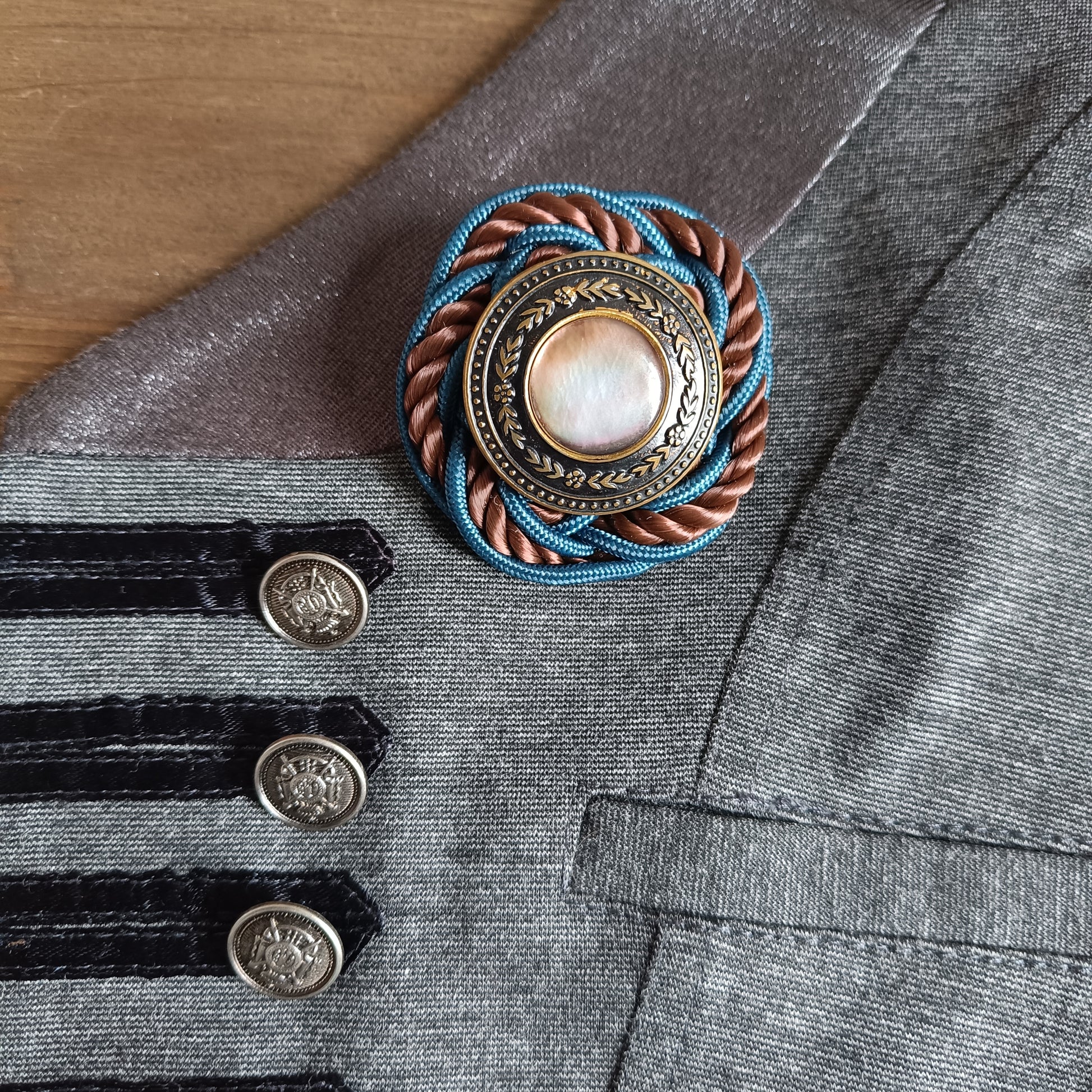 Broche ronde en passementerie avec cordon tressé bleu et brun. Au centre, un bouton vintage en métal ouvragé décoré de motifs floraux encadre un cabochon nacré irisé. La broche est épinglée sur un gilet gris