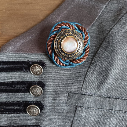 Broche ronde en passementerie avec cordon tressé bleu et brun. Au centre, un bouton vintage en métal ouvragé décoré de motifs floraux encadre un cabochon nacré irisé. La broche est épinglée sur un gilet gris