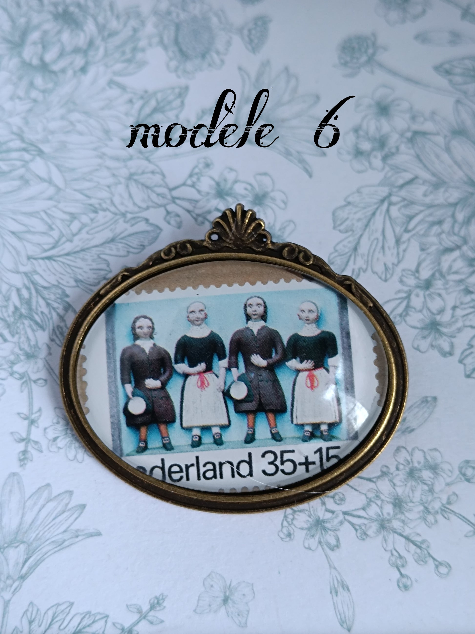 Broche ovale encadrant un timbre néerlandais vintage du siècle dernier, illustré de cinq personnages en tenue traditionnelle sur fond bleu clair, avec la mention "Nederland 35+15". Présentée sur un fond fleuri aux lignes fines et élégantes