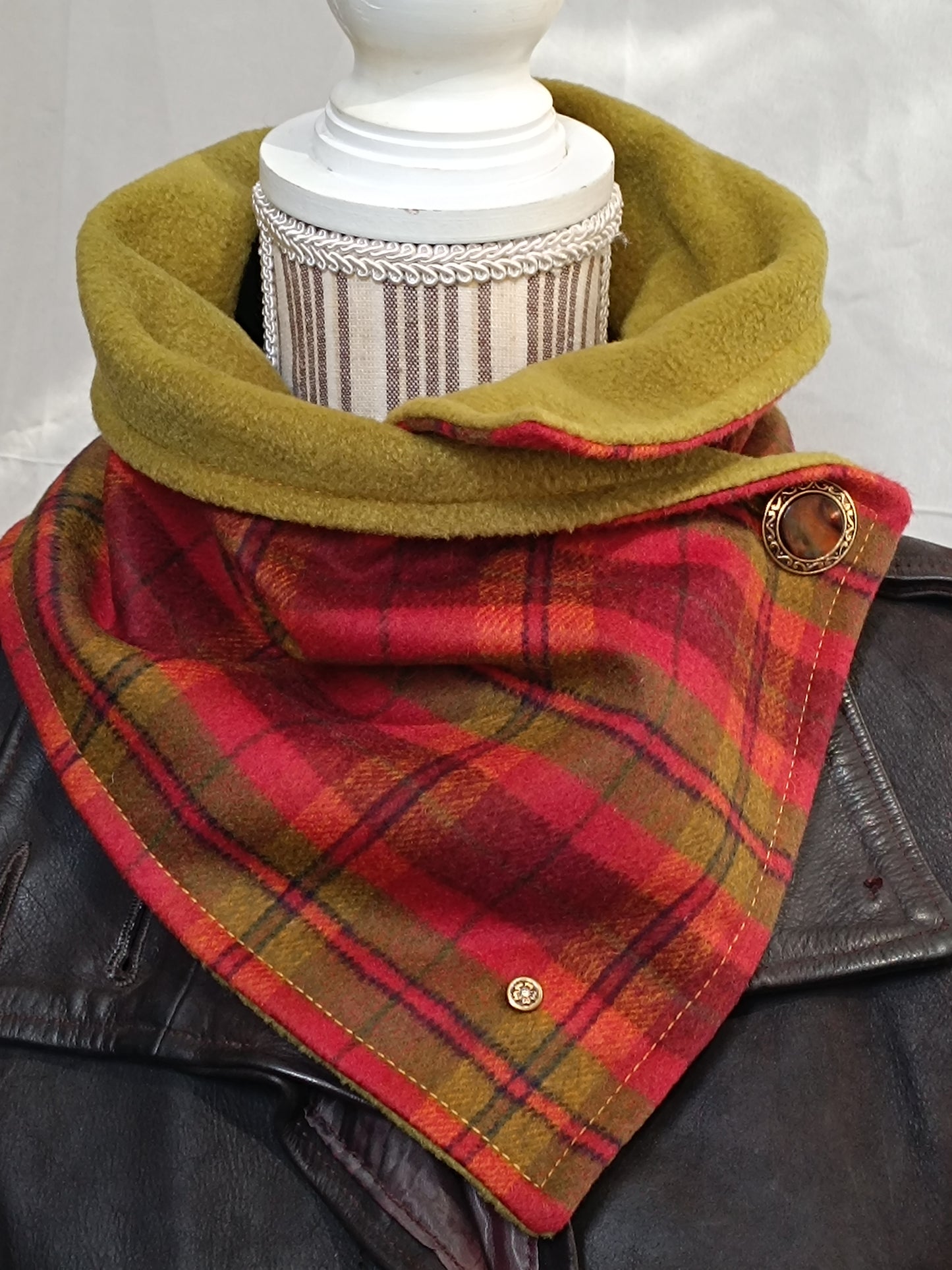 Tour de cou artisanal en laine grattée tartan rouge et vert, doublé polaire vert pistache, porté sur veste en cuir brun. Accessoire textile au style néo-bourgeois affirmé, mêlant héritage vintage revisité, féminité soignée et côté urbain. Bouton doré à motif marbré et rivet signature fleurs de cerisier visibles.