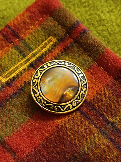 Boutonnière réalisée en fil jaune sur tartan en laine grattée rouge, orange, vert et jaune. Bouton vintage doré à centre marbré posé sur le tissu, en cours de confection. Photo d’atelier mettant en valeur le soin apporté aux finitions et le contraste lumineux des surpiqûres.