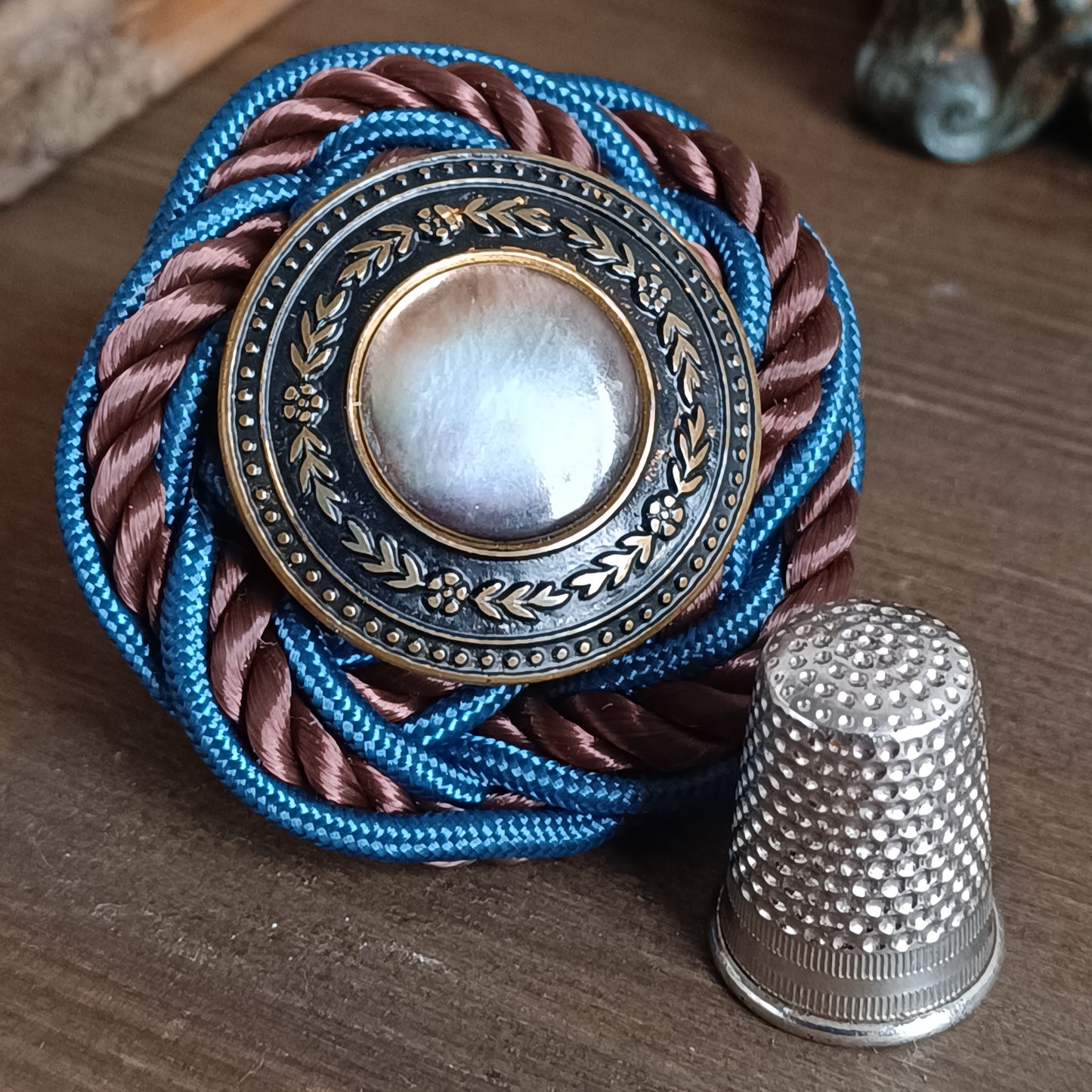 Broche ronde en passementerie avec cordon tressé bleu et brun. Au centre, un bouton vintage en métal ouvragé décoré de motifs floraux encadre un cabochon nacré irisé. La broche est posée sur une surface en bois, avec un élément sculpté flou en arrière‑plan. dimensions 