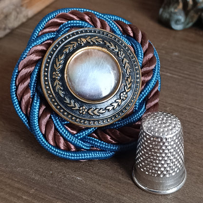 Broche ronde en passementerie avec cordon tressé bleu et brun. Au centre, un bouton vintage en métal ouvragé décoré de motifs floraux encadre un cabochon nacré irisé. La broche est posée sur une surface en bois, avec un élément sculpté flou en arrière‑plan. dimensions 