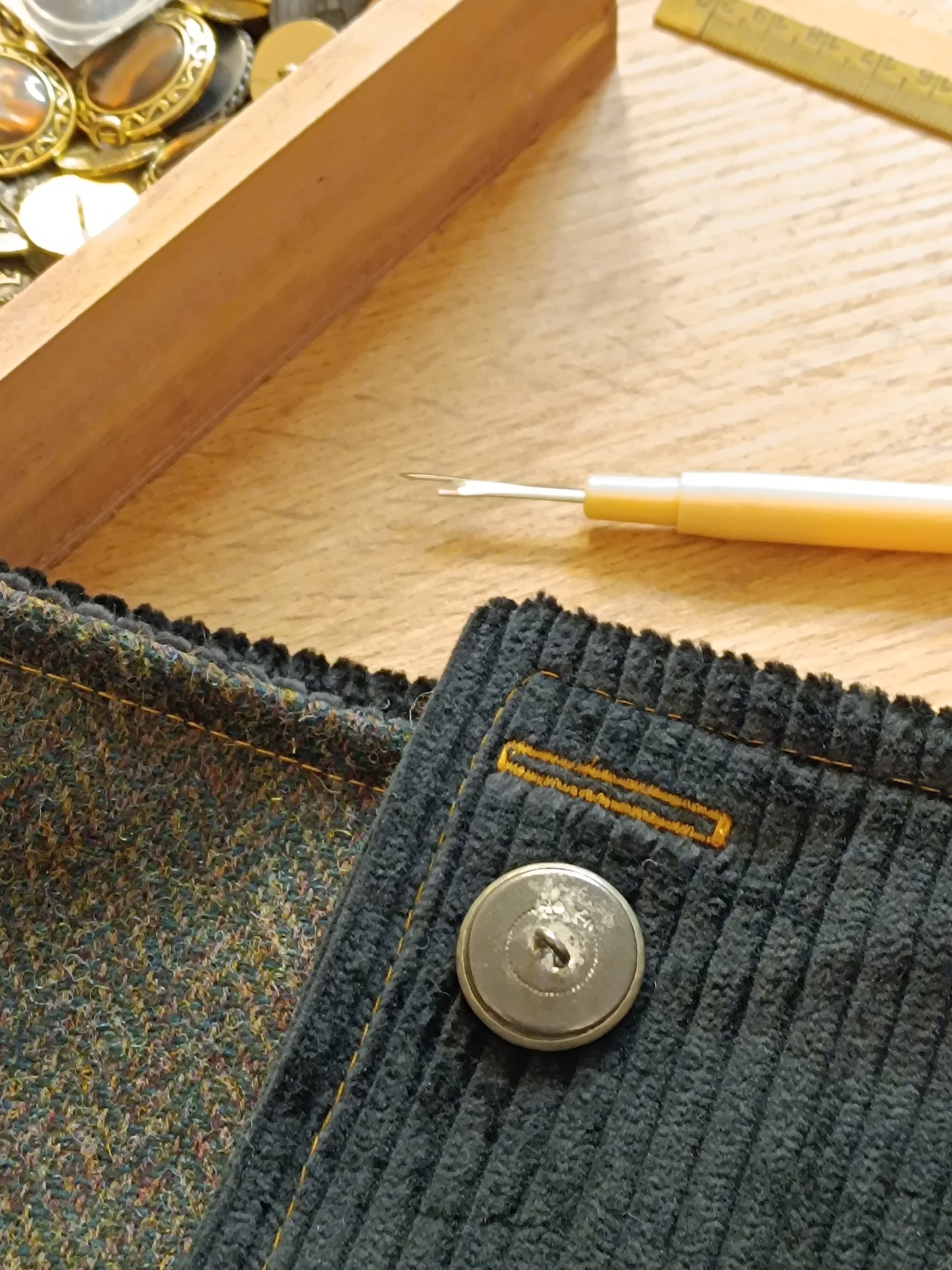 Gros plan sur bouton vintage en métal blanc vu de dos, fixé sur velours côtelé noir avec boutonnière cousue en fil jaune ocre. Ce véritable bouton ancien est issu de surcyclage, soigneusement sélectionné pour sa patine et son caractère. Détail textile artisanal, témoin du travail en cours dans notre atelier de l’Oise. Idéal pour accessoire fait main au style féerique, bohème ou médiéval.