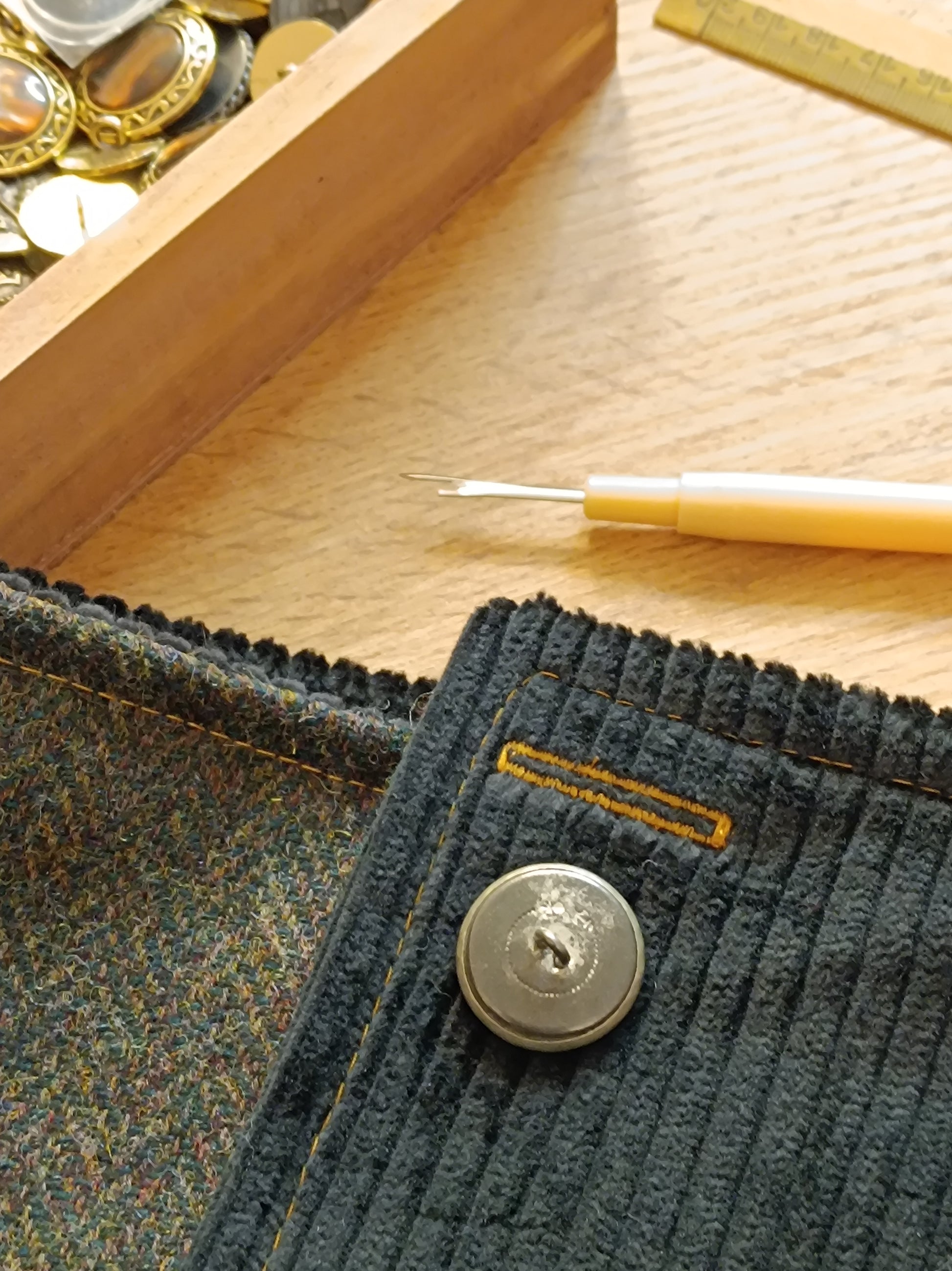 Gros plan sur bouton vintage en métal blanc vu de dos, fixé sur velours côtelé noir avec boutonnière cousue en fil jaune ocre. Ce véritable bouton ancien est issu de surcyclage, soigneusement sélectionné pour sa patine et son caractère. Détail textile artisanal, témoin du travail en cours dans notre atelier de l’Oise. Idéal pour accessoire fait main au style féerique, bohème ou médiéval.