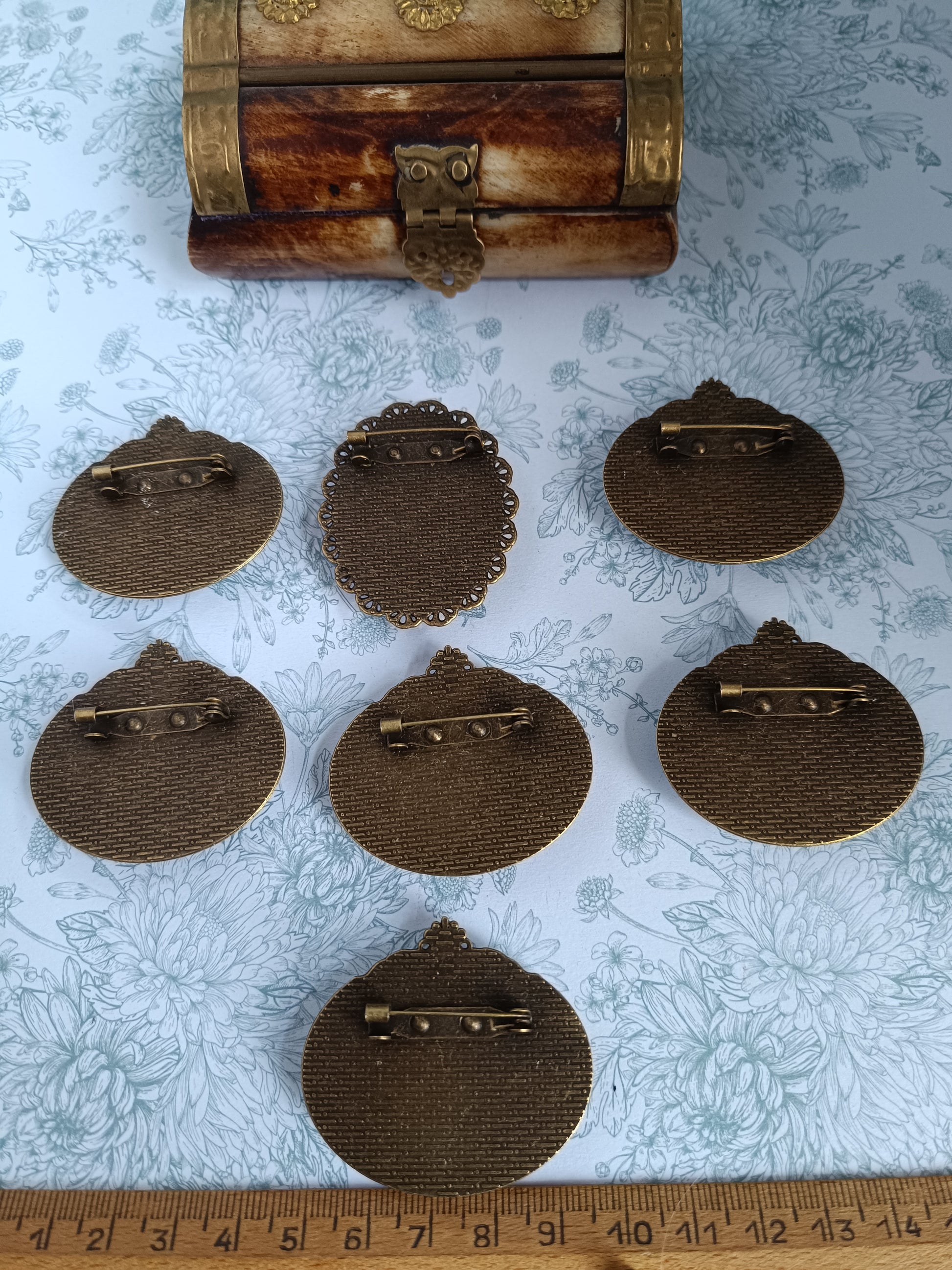 Vue de sept dos de broches vintage en métal texturé, disposés sur un fond fleuri, avec un petit coffret décoratif en laiton en haut de l’image. Une règle en bois indique un diamètre d’environ 5 cm pour chaque broche.