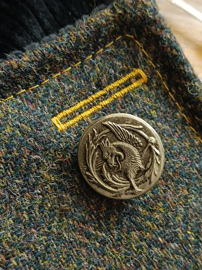 Gros plan sur la boutonnière cousue en fil jaune ocre et le bouton vintage en métal blanc orné d’un dragon flamboyant, gueule ouverte et puissance affirmée. Le lainage à chevrons révèle un subtil mélange de verts, bleus, jaunes et touches chaudes. Détail textile artisanal idéal pour accessoire fait main au style féerique, bohème ou médiéval, réalisé dans notre atelier de l’Oise.