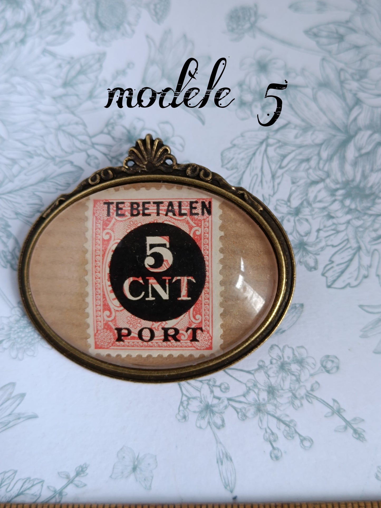 Broche ovale encadrant un timbre néerlandais vintage du siècle dernier, rouge et blanc, avec cercle noir central marqué "TE BETALEN 5 CNT PORT". Présentée sur un fond fleuri bleu pâle, dans un cadre en laiton décoratif.