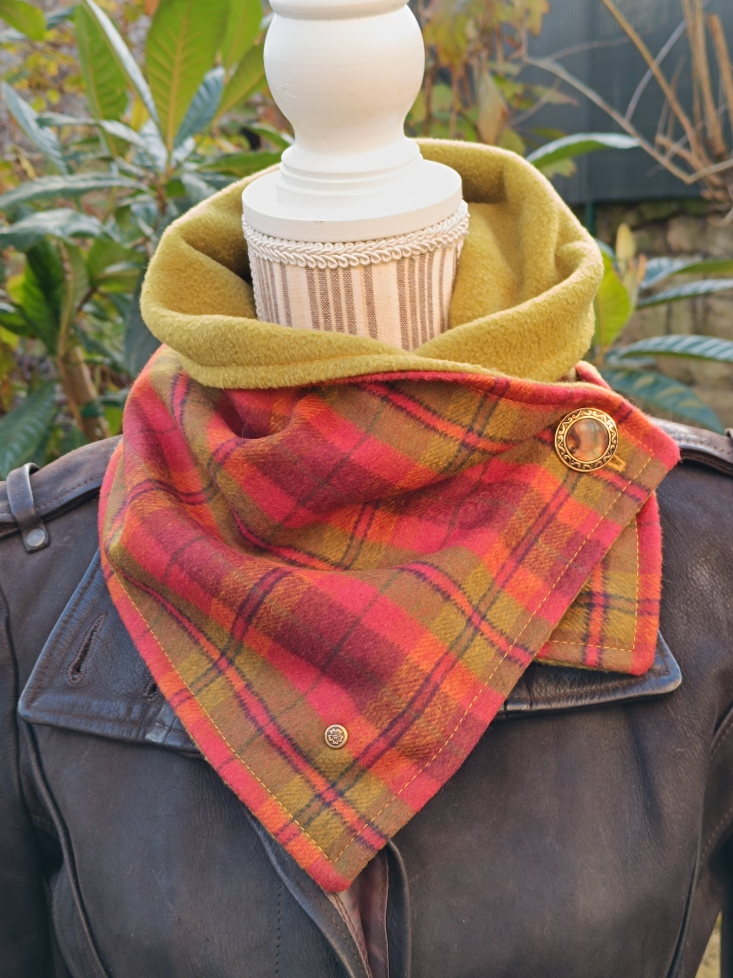 Tour de cou artisanal en tartan rouge, orange et vert aux couleurs chatoyantes, doublé polaire vert pistache, présenté sur buste de couture rayé avec veste en cuir foncé. Bouton vintage doré à motif central et rivet bronze fleurs de cerisier visibles.