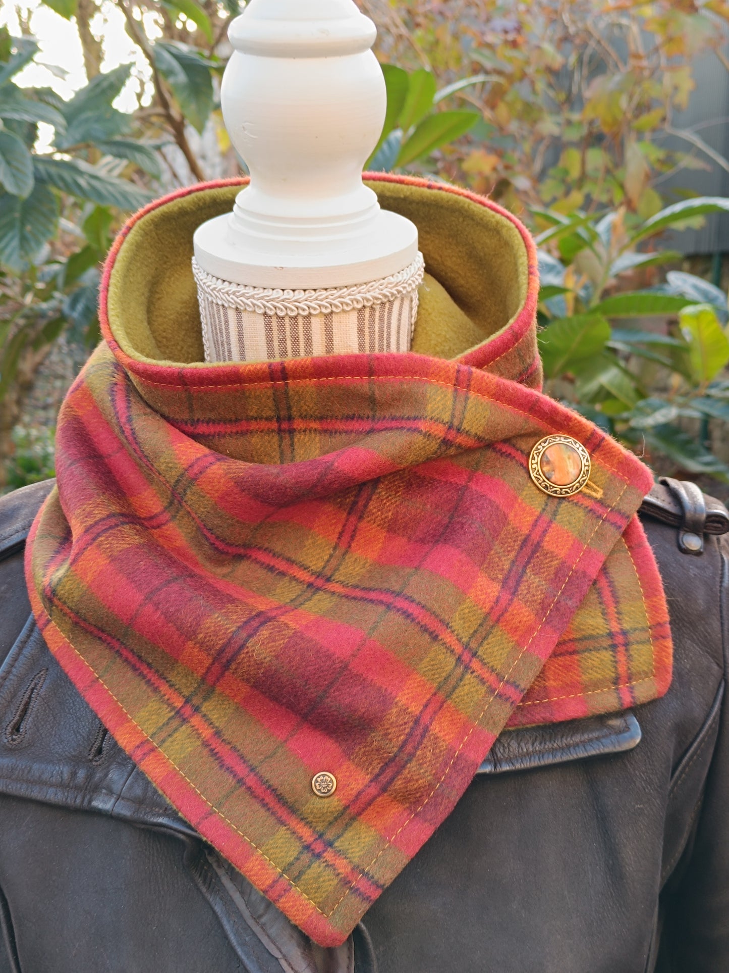 Col de bénitier tartan artisanal aux couleurs chatoyantes, doublé polaire vert pistache, porté sur veste en cuir foncé. Accessoire textile d’hiver au charme rétro et à l’élégance structurée, avec bouton vintage doré et rivet signature bronze fleurs de cerisier.