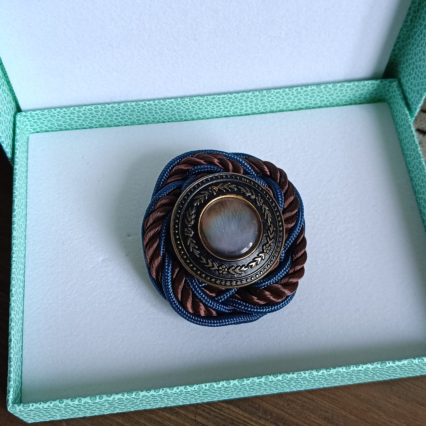 Broche ronde en passementerie avec cordon tressé bleu et brun. Au centre, un bouton vintage en métal ouvragé décoré de motifs floraux encadre un cabochon nacré irisé. dans sa boite d'envoi