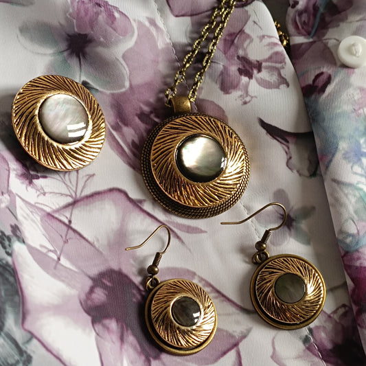 Parure de bijoux vintage en bronze antique et nacre irisée posée sur tissu fleuri clair violet pour montrer l’effet du bijou sur vêtements féminins lumineux. Ensemble artisanal rétro comprenant collier, boucles d’oreilles et broche. Le charme vintage français 
