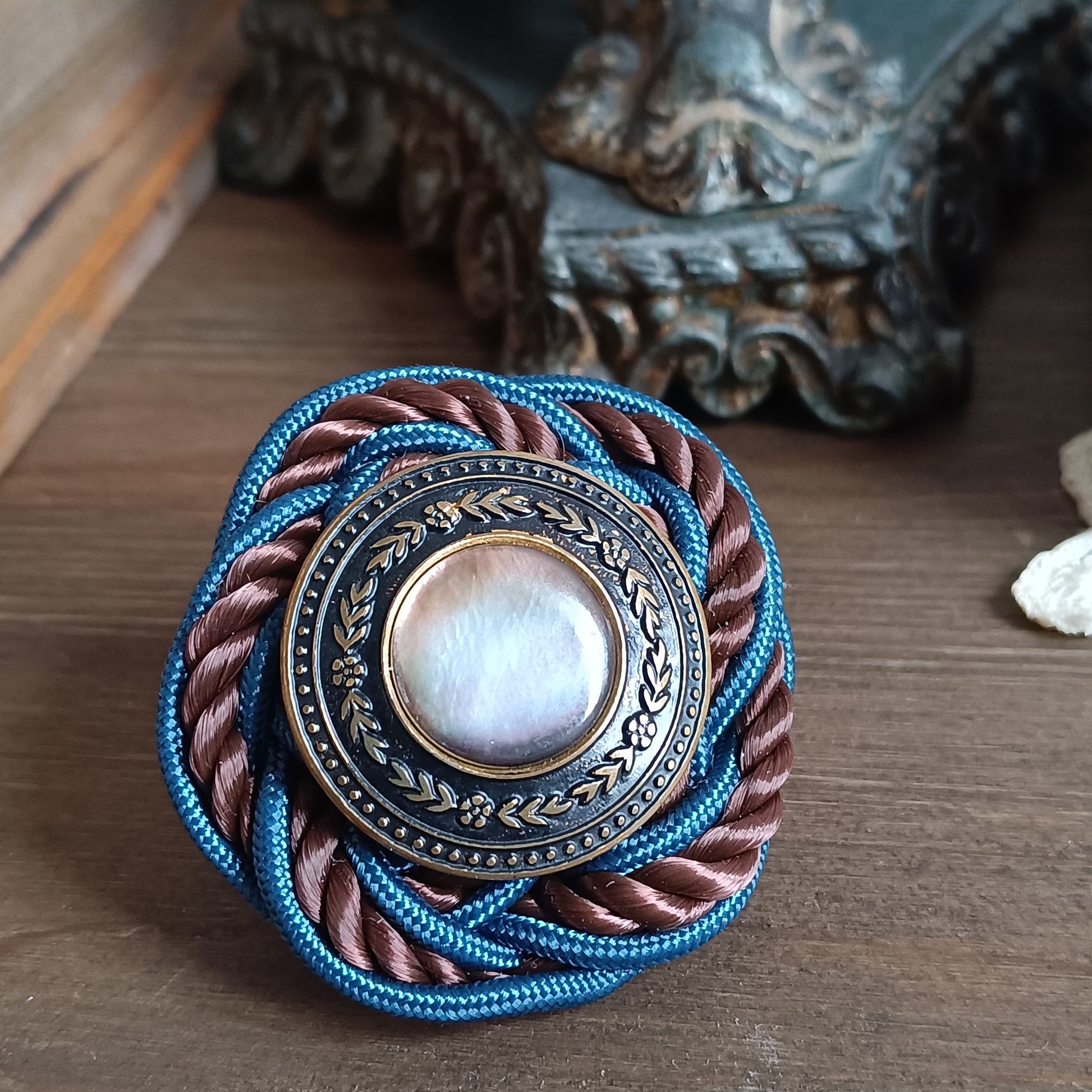 Broche ronde en passementerie avec cordon tressé bleu et brun. Au centre, un bouton vintage en métal ouvragé décoré de motifs floraux encadre un cabochon nacré irisé. La broche est posée sur une surface en bois, avec un élément sculpté flou en arrière‑plan. vue générale du bijoux 